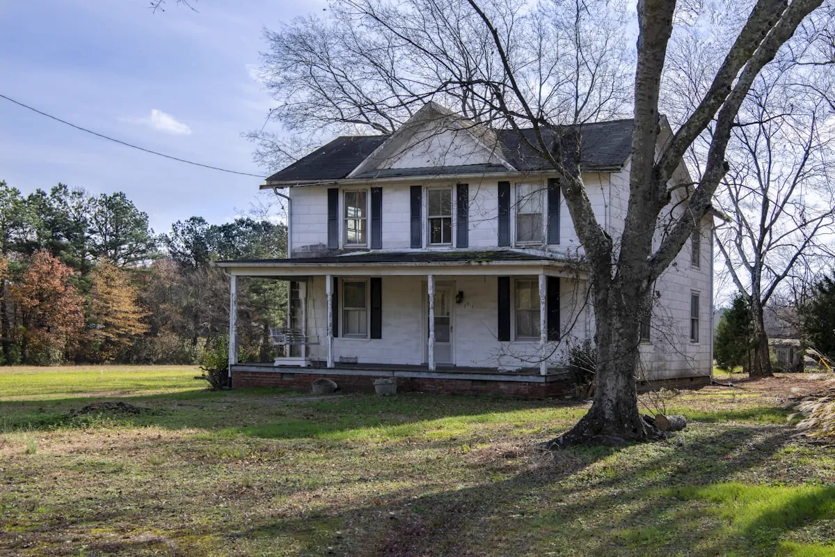 Older white two-story farmhouse with a covered front porch and a large bare tree in the yard